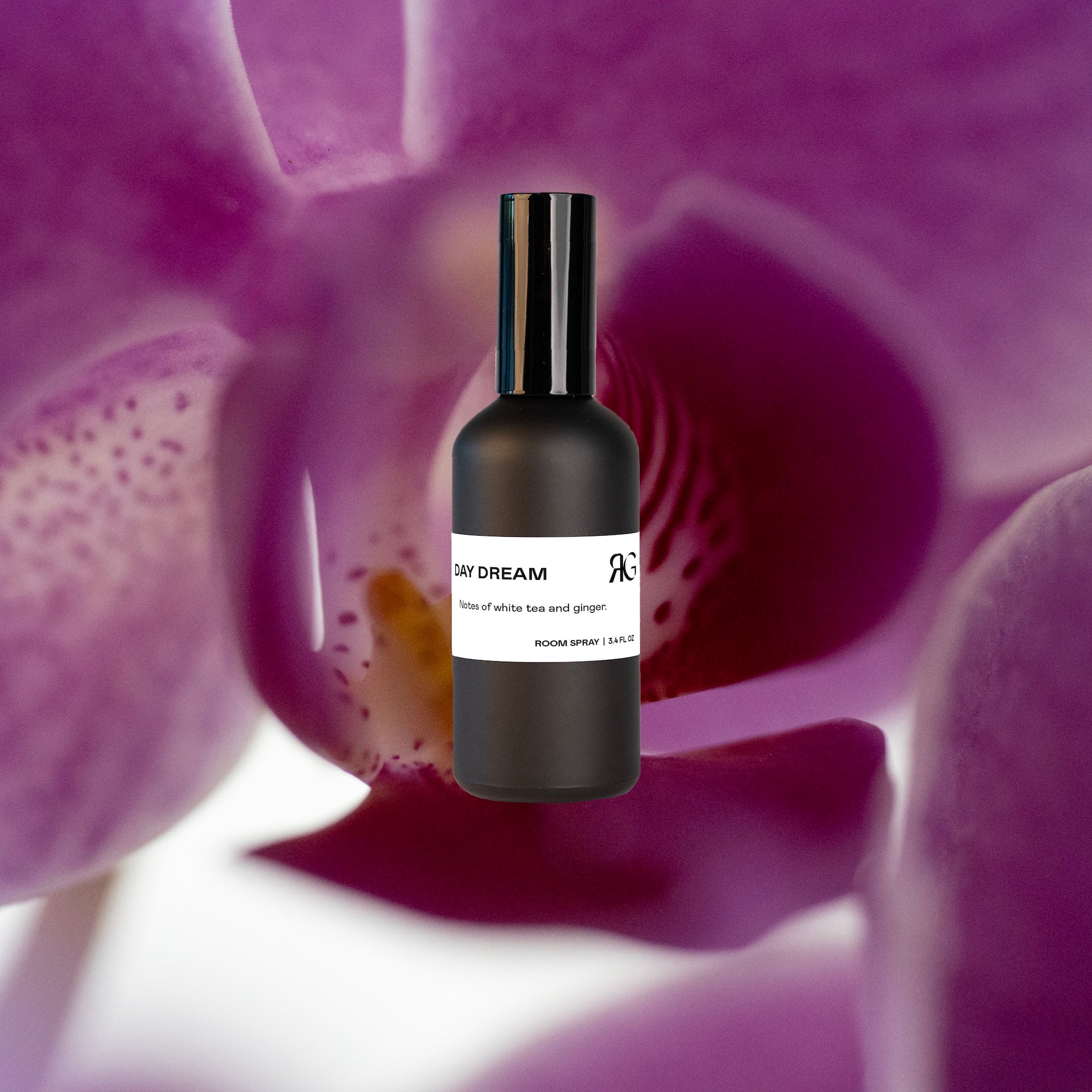 A black bottle labeled Day Dream Room Spray is centered against a blurred backdrop of purple and white petals, evoking an uplifting aroma with notes of ginger and white tea.