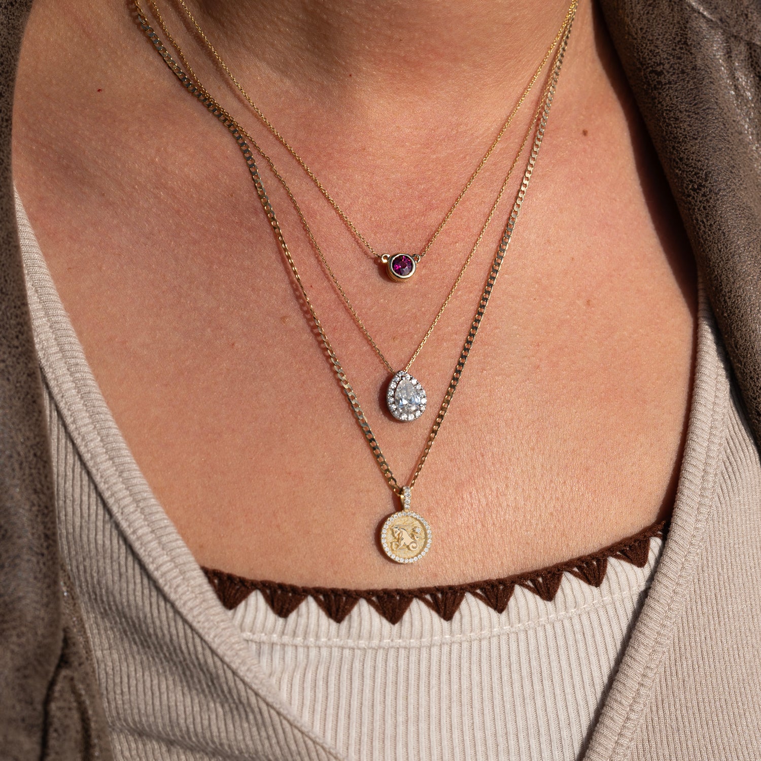 Close-up of a person wearing three layered 14k gold necklaces, including the 14k Garnet Bezel Necklace with an ethically sourced garnet, a teardrop pendant necklace, and a coin pendant necklace over a beige ribbed top with brown scalloped edges.