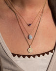 Close-up of a person wearing three layered 14k gold necklaces, including the 14k Garnet Bezel Necklace with an ethically sourced garnet, a teardrop pendant necklace, and a coin pendant necklace over a beige ribbed top with brown scalloped edges.