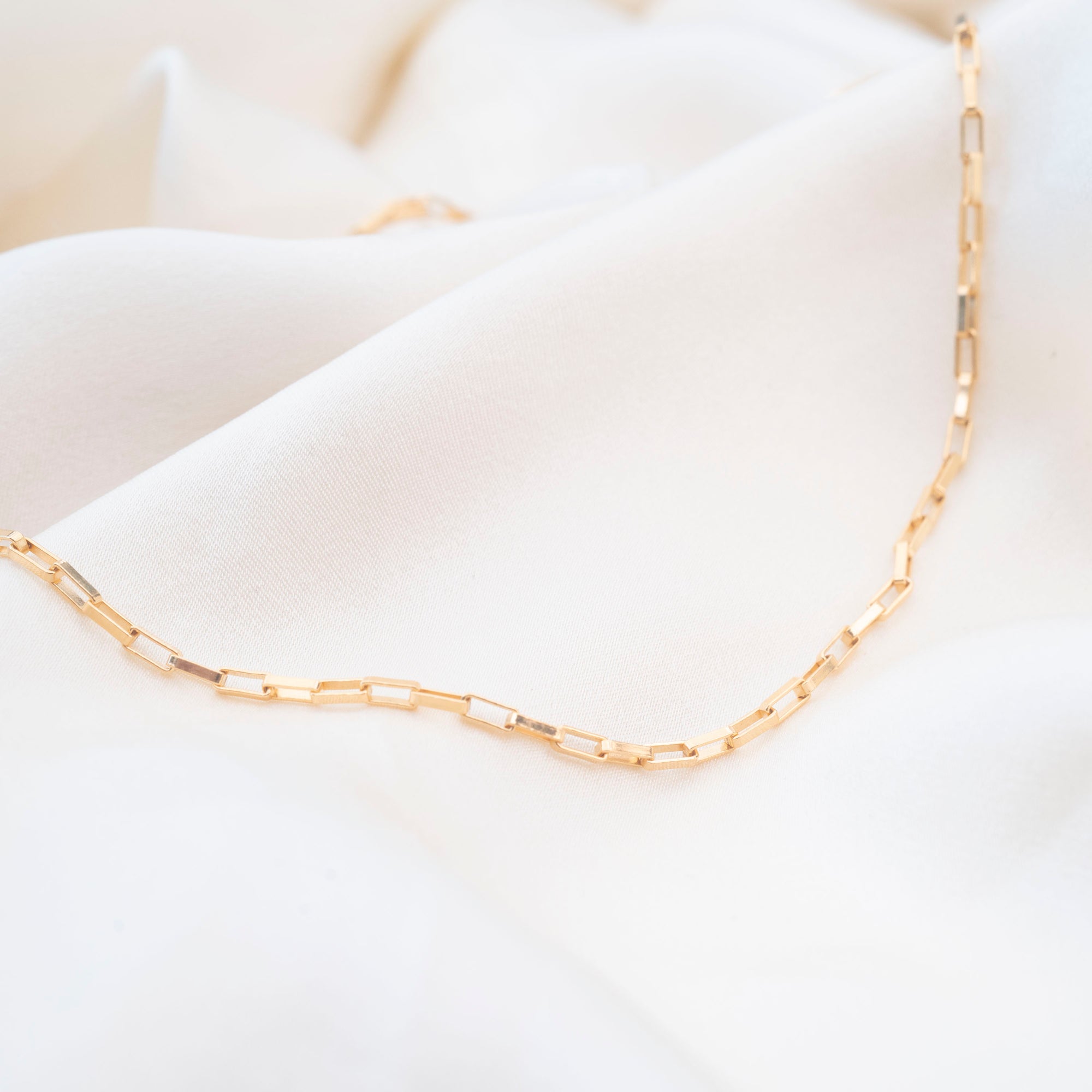 The Gold Filled Box Necklace features rectangular links and rests on soft white fabric, offering an elegant and timeless look.