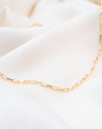 The Gold Filled Box Necklace features rectangular links and rests on soft white fabric, offering an elegant and timeless look.