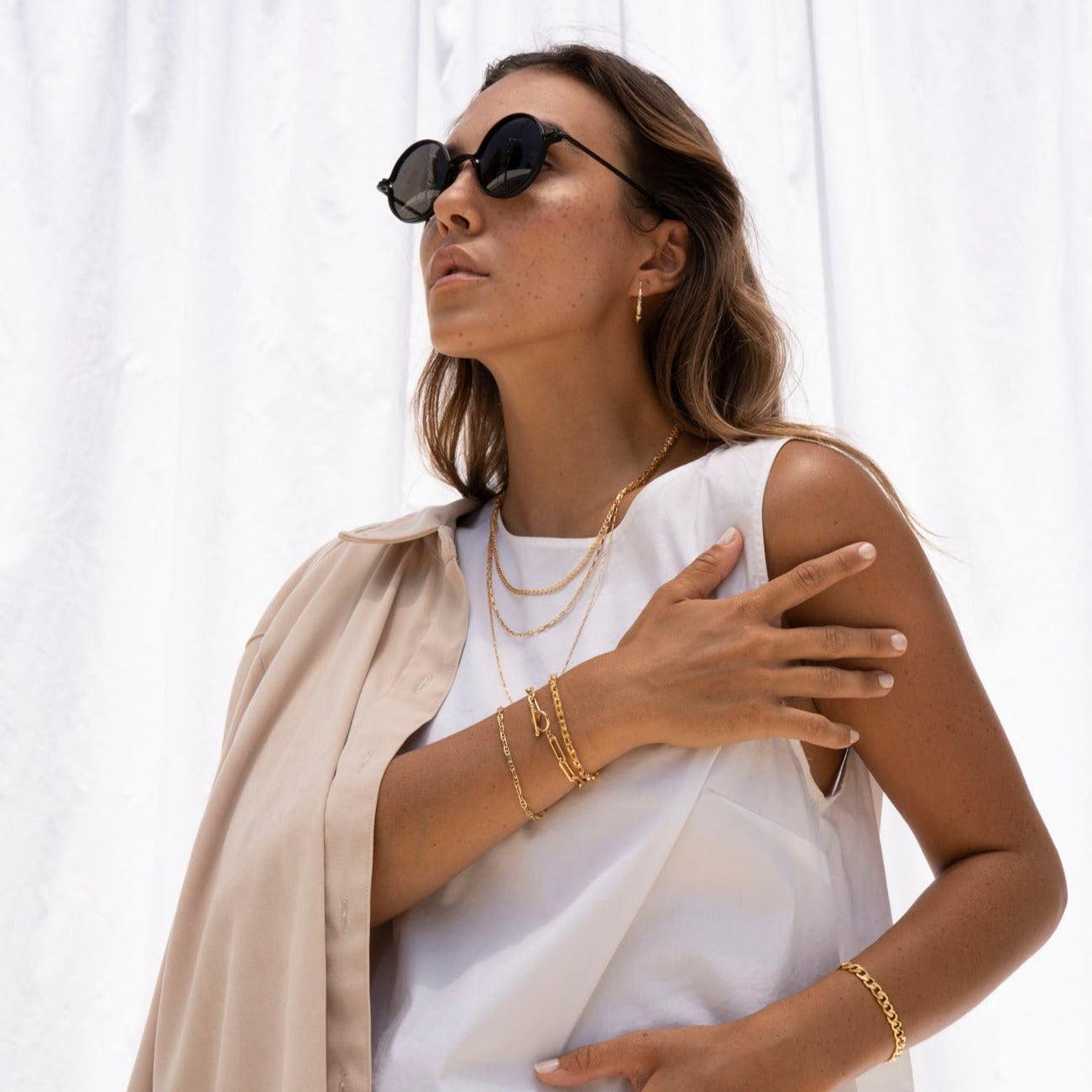 A woman in sunglasses, gold jewelry—including a Gold Filled Box Necklace—and a white sleeveless top poses with one hand on her shoulder, a beige jacket draped over her arm. She stands in front of a white backdrop.