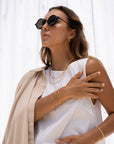 A woman in sunglasses, gold jewelry—including a Gold Filled Box Necklace—and a white sleeveless top poses with one hand on her shoulder, a beige jacket draped over her arm. She stands in front of a white backdrop.