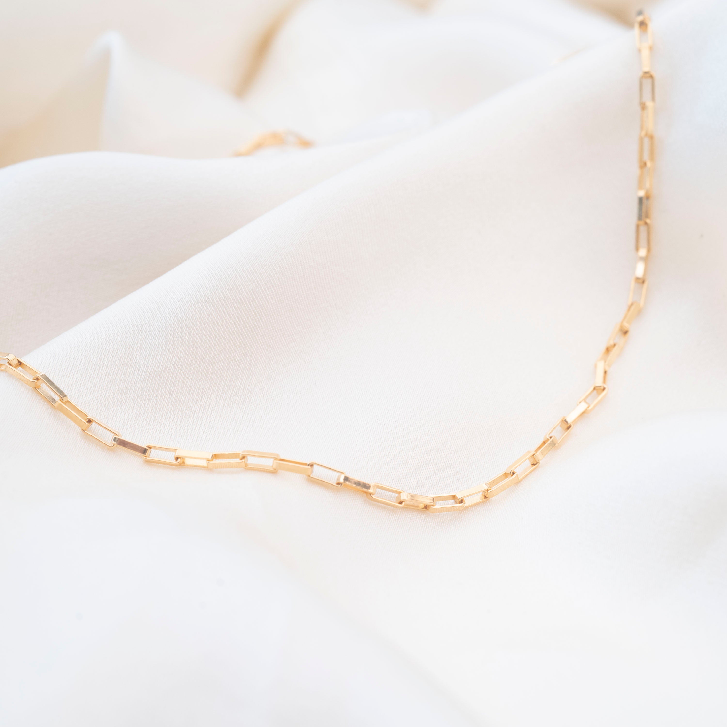 The Gold Filled Box Necklace features rectangular links and rests on soft white fabric, offering an elegant and timeless look.